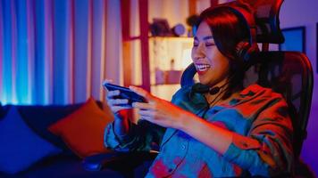 Happy asia girl gamer wear headphone competition play video game online with smartphone colorful neon lights in living room at home. Esport streaming game online, Home quarantine activity concept. photo