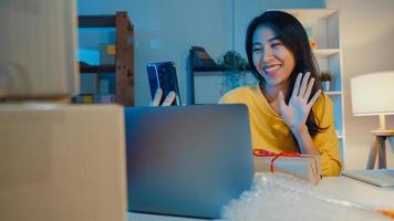 Una joven empresaria asiática usa un teléfono inteligente para recibir una orden de compra y mostrar el empaque del producto al cliente, en vivo, en línea, en la tienda por la noche. propietario de una pequeña empresa, concepto de entrega de mercado en línea.