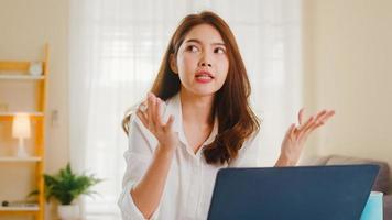 Asian businesswoman using laptop talk to colleagues about plan in video call while smart working from home at living room. Self-isolation, social distancing, quarantine for coronavirus prevention. photo