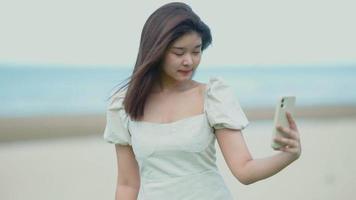 linda garota asiática caminhando na praia, ela usava o celular para passear tirando fotos de selfies video