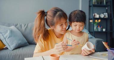 Happy cheerful Asia family mom teach toddler girl paint ceramic pot having fun relax on table in living room at house. Spending time together, Social distance, Quarantine for coronavirus prevention. photo