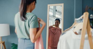 Beautiful attractive Asia lady choosing clothes on clothes rack dressing looking herself in mirror in living room at house. Girl think what to wear casual shirt. Lifestyle women relax at home concept. photo