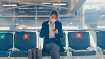 Asian business lady traveller wear suit sitting with suitcase and use smart phone chat message in bench wait for flight at airport. Business travel commuter in covid pandemic, Business travel concept. photo