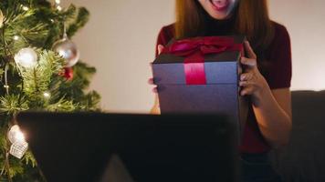 Mujer joven de Asia con videollamada de tableta hablando con pareja con caja de regalo de Navidad, árbol de Navidad decorado con adornos en la sala de estar en casa. noche de navidad y fiesta de año nuevo.