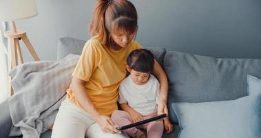 Happy cheerful Asia family mom and cute kid using digital tablet interest cartoon and watch movie having fun relax on couch in living room at house. Spending time together, Quarantine for coronavirus. photo