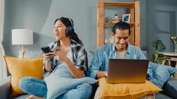 Happy young asian couple activity man use laptop computer work relax enjoy with women wear headphones use smartphone listen music at sofa in living room at house. Young married work from home concept. photo