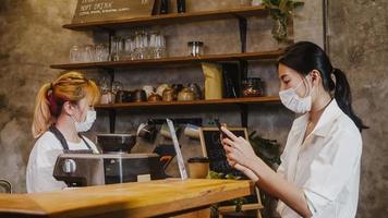 Young Asia female wear face mask self service use mobile phone pay contactless at restaurant. Girl barista talking receive order from customer at cafe. Lifestyle new normal after corona virus concept. photo