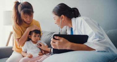 Young Asia female pediatrician hold stethoscope exam little girl patient visit doctor with mother sit on couch in living room at house. Medical care insurance, Visit patient at home concept. photo
