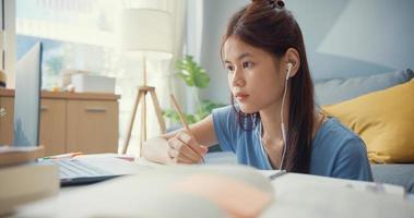 joven adolescente de asia con ropa casual auriculares usar computadora portátil aprender en línea escribir cuaderno de conferencias en la sala de estar en casa. Aislar el concepto de pandemia de coronavirus de educación en línea e-learning. foto