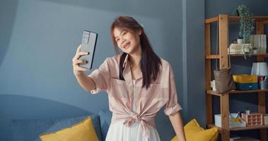 Happy carefree young Asia girl use smartphone video call enjoy talk with college friend in living room at home. Social distance coronavirus pandemic concept. Freedom and active lifestyle concept. photo