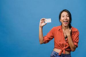 Joven mujer asiática muestra una tarjeta bancaria de crédito con expresión positiva, sonríe ampliamente, vestida con ropa informal sintiendo felicidad y parada aislada sobre fondo azul. concepto de expresión facial. foto