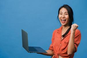 Young Asia lady using laptop with positive expression, smiles broadly, dressed in casual clothing feeling happiness and stand isolated on blue background. Happy adorable glad woman rejoices success. photo