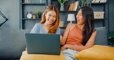 Two Asia lesbian women site on couch together looking at laptop screen in living room at home together. Happy couple roommate ladies enjoy web surfing online shopping, Lifestyle woman at home concept.
