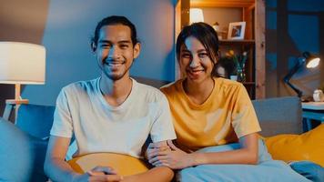 Feliz pareja de jóvenes asiáticos hombre y mujer mirando a la cámara sonríen y alegres en la videollamada en línea por la noche en la sala de estar en casa, quedarse en casa en cuarentena, vida matrimonial, concepto de distanciamiento social.