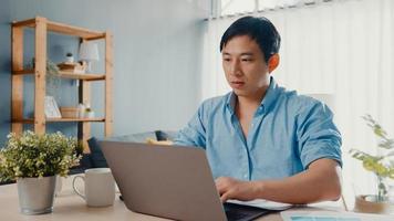 Freelance Asia guy casual wear using laptop online learning in living room at house. Working from home, remotely work, distance education, social distancing, quarantine for corona virus prevention. photo