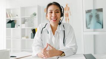 Young Asia lady doctor in white medical uniform with stethoscope using computer laptop talk video conference call with patient, looking at camera in health hospital. Consulting and therapy concept. photo