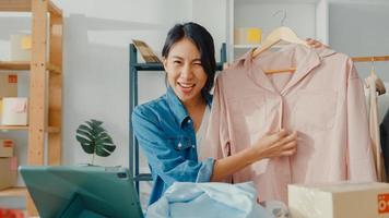 Young Asia lady fashion designer using mobile phone receiving purchase order and showing clothes recording video live streaming online at shop. Small business owner, online market delivery concept. photo