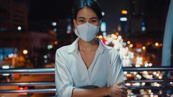 Exitosa empresaria asiática joven en ropa de oficina de moda usa mascarilla médica sonriendo y mirando a la cámara mientras feliz está sola al aire libre en la noche urbana de la ciudad moderna. concepto de negocio en marcha.