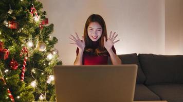 Young Asia female using laptop video call talking with couple with X'Mas present box, Christmas tree decorated with ornament in living room at home. Christmas night and New Year holiday festival.
