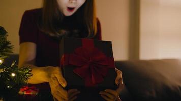 Joven mujer asiática divirtiéndose abriendo caja de regalo de Navidad cerca del árbol de Navidad decorado con adornos en la sala de estar en casa. Feliz noche de navidad y feliz año nuevo festival navideño. foto