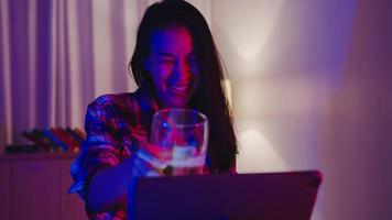 Young Asia lady drinking beer having fun happy moment disco neon night party event online celebration via video call in living room at home. Social distancing, quarantine for coronavirus prevention. photo