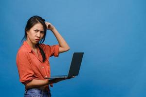 Young Asia lady using laptop with negative expression, excited screaming, cry emotional angry in casual cloth and stand isolated on blue background with blank copy space. Facial expression concept. photo