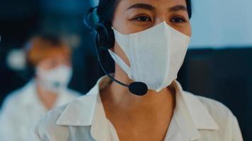 Millennial Asia young call center team or customer support service executive wearing face mask prevent covid-19 using computer and microphone headset working technical support in late night office. photo