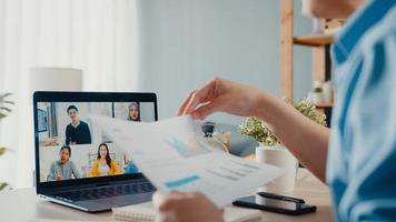 Young Asia businessman using laptop talk to colleagues about plan in video call meeting while work from home at living room. Self-isolation, social distancing, quarantine for corona virus prevention. photo