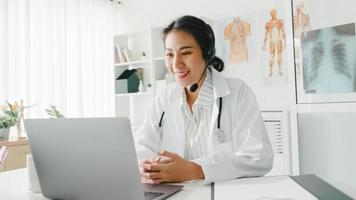 Joven doctora asiática en uniforme médico blanco con estetoscopio usando computadora portátil hablando por videoconferencia con el paciente en el escritorio en la clínica de salud u hospital. concepto de consulta y terapia. foto