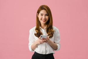 Surprised young Asia lady using mobile phone with positive expression, smiles broadly, dressed in casual clothing and looking at camera on pink background. Happy adorable glad woman rejoices success. photo