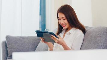 Mujer de negocios asiática joven que usa la videollamada de la tableta que habla con la familia mientras trabaja desde casa en la sala de estar. autoaislamiento, distanciamiento social, cuarentena por coronavirus en el próximo concepto normal. foto
