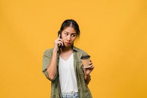 Jovencita asiática habla por teléfono y sostiene la taza de café con expresión negativa, gritos emocionados, llora enojado emocionalmente en ropa casual y se para aislado sobre fondo amarillo. concepto de expresión facial. foto