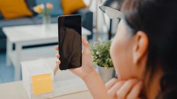 La jovencita asiática usa un teléfono inteligente con pantalla negra en blanco simulada para mostrar texto publicitario mientras trabaja de manera inteligente desde casa en la sala de estar. tecnología chroma key, concepto de diseño de marketing. foto
