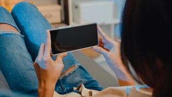 Joven mujer asiática usa un teléfono inteligente con pantalla negra en blanco simulada para mostrar texto publicitario mientras descansa en el sofá de la sala de estar en la moderna noche de casa. tecnología chroma key, concepto de diseño de marketing.