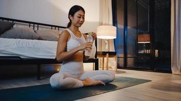 Young Asia lady doing yoga exercise working out and drinking pure water in living room at home night. Sport and recreation activity, social distancing, quarantine for corona virus prevention concept. photo