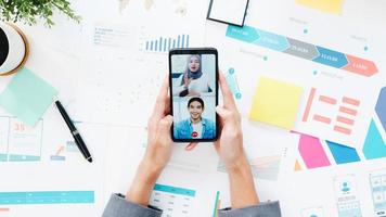 Top view of young Asia businesswoman using phone talk to colleague about plan in video call meeting while work from home at living room. Social distancing, quarantine for corona virus prevention. photo