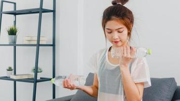 Young Korean lady in sportswear exercises doing working out using light weight dumbbell with bottle of water on couch in living room at home. Social distance, Isolation during the virus, stay at home. photo