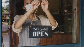 Young Asia girl wear face mask turning a sign from closed to open sign on door looking outside waiting for clients after lockdown. Owner small business, food and drink, business reopen again concept. photo