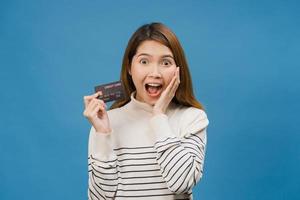 Joven mujer asiática muestra una tarjeta bancaria de crédito con expresión positiva, sonríe ampliamente, vestida con ropa informal sintiendo felicidad y parada aislada sobre fondo azul. concepto de expresión facial. foto