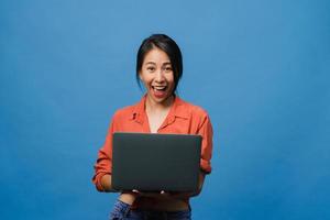 Surprised young Asia lady using laptop with positive expression, smile broadly, dressed in casual clothing and looking at camera on blue background. Happy adorable glad woman rejoices success. photo