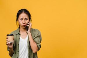 Young Asia lady talk by phone and hold coffee cup with negative expression, excited screaming, cry emotional angry in casual cloth and stand isolated on yellow background. Facial expression concept. photo