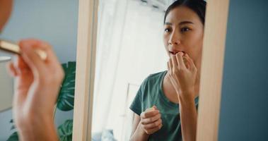 Beautiful Asia lady with casual cloth put lipstick on her lips in front of mirror in bedroom at home in morning before go dating outside. Smiling young woman applying makeup and looking at mirror. photo