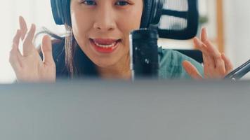 feliz chica asiática graba un podcast en su computadora portátil con auriculares y micrófono habla con la audiencia en su habitación. Podcaster femenina hace podcast de audio desde su estudio en casa, permanece en el concepto de casa.