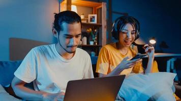 Freelance asia couple man and woman in casual hasband work  laptop computer at night enjoy happy relax with wife wear headphone listen music in smartphone sing dance on couch at house, Married life. photo
