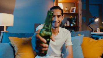 Happy young asia man looking at camera enjoy night party event online with friends toast drink beer via video call online in living room at home, Stay at home quarantine, Social distancing concept. photo