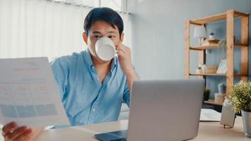 freelance asia guy casual wear usando laptop y tomando café en la sala de estar en casa. trabajo desde casa, trabajo a distancia, educación a distancia, distanciamiento social, cuarentena para la prevención del coronavirus. foto