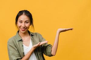 Portrait of young Asian lady smiling with cheerful expression, shows something amazing at blank space in casual cloth and looking at camera isolated over yellow background. Facial expression concept. photo