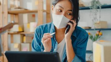 Young Asia businesswomen wear face mask using mobile phone call receiving purchase order and check product on stock work at home office. Small business owner, online market delivery freelance concept. photo