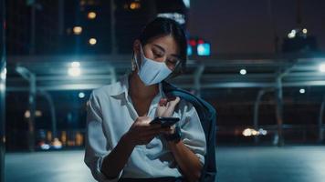 Young Asia businesswoman in fashion clothes wearing face mask using smart phone typing text message while stand outdoor in urban city at night. Social distancing to prevent spread of COVID-19 concept. photo