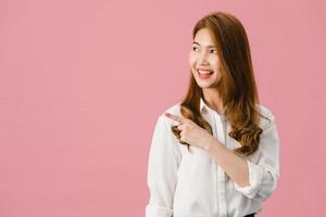 Retrato de joven asiática sonriendo con expresión alegre, muestra algo sorprendente en el espacio en blanco en ropa casual y de pie aislado sobre fondo rosa. concepto de expresión facial. foto